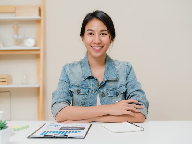 Asia business woman feeling happy smiling and looking to camera while relax at home office. Young asian woman working writing notebook document finance on desk in living room at home.